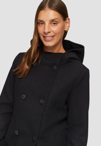 Zwart, dubbelzijdig jasje met een capuchon, met stofstructuur en knoopdetails, tentoongesteld op een model tegen een neutrale achtergrond.