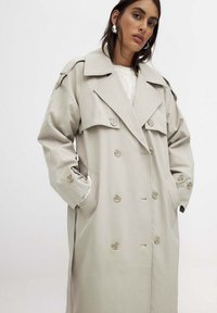 Gabardina beige con un diseño de doble botonadura, gran collar, bolsillos laterales y puños abotonados, fabricada con un material suave y ligero.