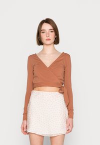 Blusa castanha de manga longa em estilo cruzado com detalhe de laço, combinada com uma saia branca com pequenos padrões florais em rosa. Tecido suave com uma silhueta ajustada.