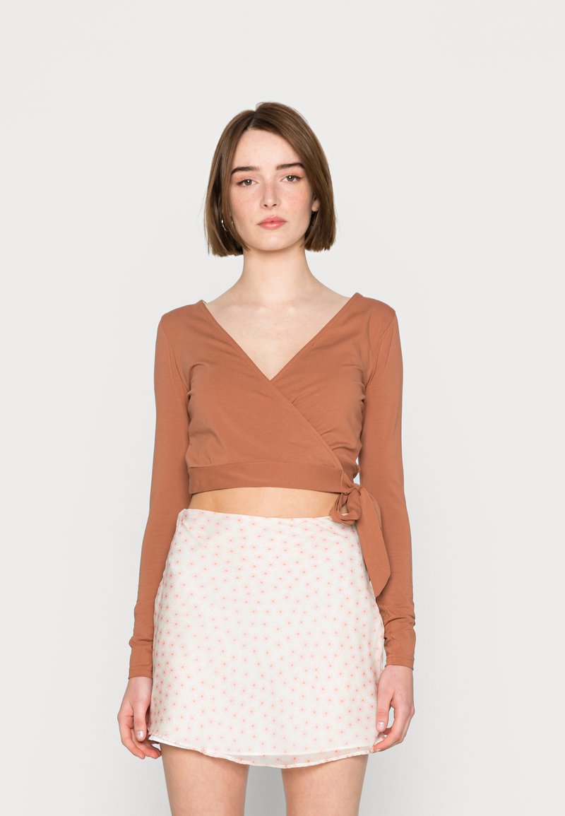 Blusa castanha de manga longa em estilo cruzado com detalhe de laço, combinada com uma saia branca com pequenos padrões florais em rosa. Tecido suave com uma silhueta ajustada.