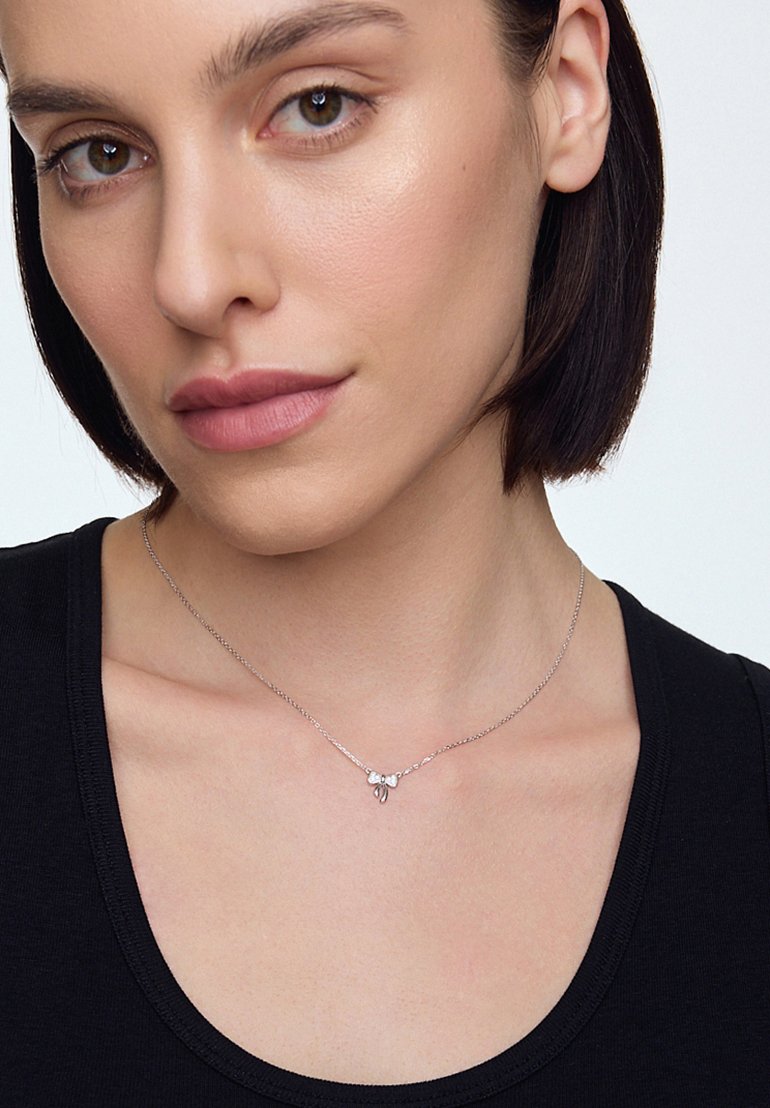 Srebrny naszyjnik z delikatnym łańcuszkiem, ozdobiony małym uroczym motylkiem oraz trzema maleńkimi kamieniami szlachetnymi. Modelka nosi czarną bluzkę.