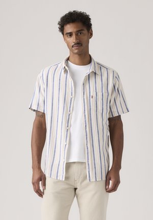 Homme portant une chemise à manches courtes beige à rayures verticales bleues par-dessus un t-shirt blanc et un pantalon de couleur claire, debout de manière neutre devant un fond uni.