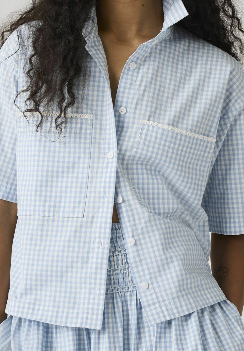Chemise à manches courtes boutonnée à motif vichy bleu et blanc avec deux poches avant, portée avec une jupe assortie à taille élastique.