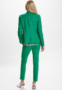 Zielony garnitur szyty na miarę z dopasowaną marynarką i wąskimi spodniami. Gładki materiał, rozcięcie z tyłu marynarki, subtelny wzór w kratkę na koszuli pod spodem.