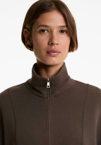 Junge Person mit kurzen braunen Haaren, die eine braune Jacke mit hohem Kragen und Reißverschluss trägt, mit neutralem Ausdruck, fotografiert vor einem schlichten weißen Hintergrund.