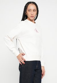 Biela mikina s rebrovanými manžetami a lemom, s malým ružovým logom na hrudi. Materiál má hladkú textúru a voľný strih.