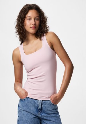 Fiatal nő göndör sötét hajjal, világos rózsaszín ujjatlan felsőt és kék farmert visel, kezét a zsebében tartva, egyszínű háttér előtt áll.