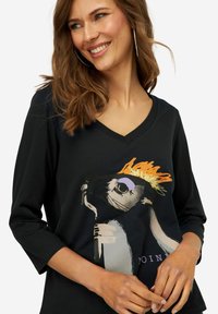 Maglietta nera a maniche lunghe con scollo a V, caratterizzata da un grafiche colorato di un animale stilizzato con una corona e la scritta "UNISCITI." Tessuto morbido.