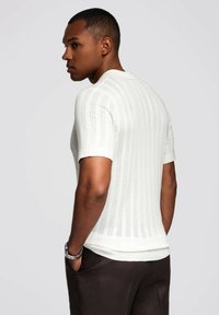 Suéter de punto blanco de manga corta con rayas verticales, ajuste slim y cuello redondo. Combinado con pantalones marrones oscuros.