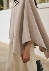 Lys beige poncho lavet af blødt strikket stof, der har et løst, draperet design med ribbet halsudskæring og kanter. Båret over en cremefarvet kjole.