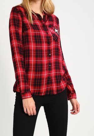 Žena nosící červenou a černou kostkovanou košili na knoflíky s dlouhým rukávem a černé kalhoty, stojící před jednobarevným pozadím.