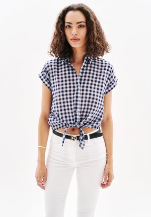 Chemise à manches courtes en vichy bleu marine et blanc avec un nœud à l'avant, associée à un pantalon blanc et une ceinture noire avec une accent en or.
