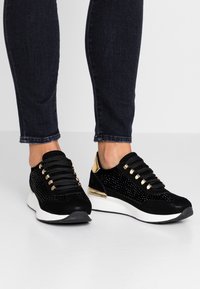 Svarta mockaskor med metalliska guldackcenter, nätdetaljer och platta snören; rund tå och vit sula för en modern atletisk look.