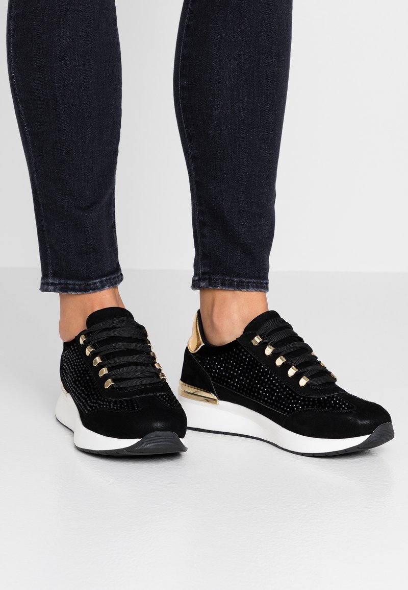 Svarta mockaskor med metalliska guldackcenter, nätdetaljer och platta snören; rund tå och vit sula för en modern atletisk look.