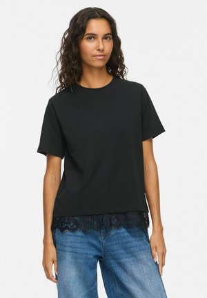 Femme aux longs cheveux bouclés portant un t-shirt noir à manches courtes avec un ourlet en dentelle et un jean bleu, debout devant un fond blanc uni.
