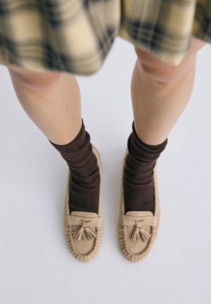 Jambes vêtues d'une jupe à carreaux beige, de chaussettes marron foncé et de mocassins beiges avec des pompons sur un fond clair.