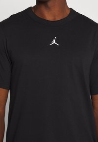 Schwarzes T-Shirt mit kurzen Ärmeln und einem kleinen weißen Jumpman-Logo, das zentral auf der Brust platziert ist, getragen von einer Person mit dunkler Hautfarbe.