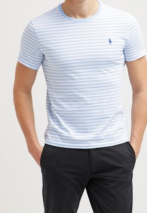 Homme portant un t-shirt à manches courtes rayé bleu clair et blanc avec un logo bleu foncé sur la poitrine, associé à un pantalon noir, mains dans les poches.