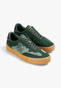 Zapatillas de ante verde con suela de goma, que presentan un bordado de calavera y huesos cruzados, cordones redondos y una textura suave en la parte superior.
