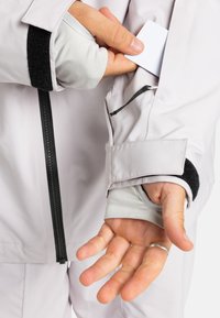 Hand, die eine hellgraue Jacke mit Reißverschluss und Klettverschluss trägt, steckt eine weiße Karte in die seitliche Tasche, während ein sichtbarer silberner Ring am kleinen Finger zu sehen ist.