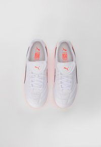 Pari valkoisia Puma Liga jalkapallokenkiä, joissa on oranssi Puma-logo ja mustat yksityiskohdat, nähtynä ylhäältä valkoista taustaa vasten.