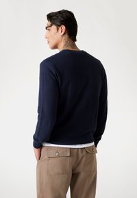 Navy blue knit sweater with textured pattern and ribbed cuffs, combined with beige pants that have two back pockets and button details.