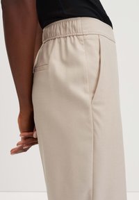 Close-up van een persoon in beige broek met een elastische tailleband en zijzakken, met de handen gekruist achter hun rug.