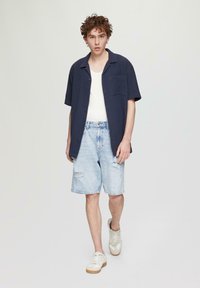 Marineblauw kortemouw shirt met knopen en een zak, witte tanktop, lichtblauwe versleten denim shorts en beige sneakers met witte accenten.