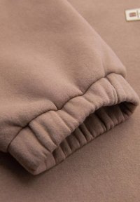 Hellbraunes Sweatshirtmaterial mit einem gerippten Bündchen. Weiche Textur, elastischer Saum und dezentes Branding in einem quadratischen Etikett.