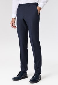 Dunkelblaue Hose in Slim-Fit mit flacher Front, Seitentaschen und einem glatten, strukturierten Stoff. Kombiniert mit dunkelbraunen Lederschuhen.
