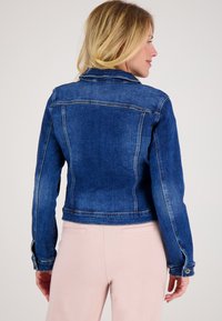 Vrouw die zijwaarts kijkt, gekleed in een strakke blauwe denimjack en lichtroze broek, tegen een effen achtergrond.