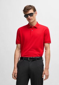 Röd polo skjorta med krage och korta ärmar, strukturerad tyg, tillsammans med mörka byxor och ett svart bälte. Svarta solglasögon fulländar looken.