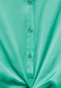 Camicia abbottonata turchese in tessuto morbido, con una placca centrale e cinque bottoni coordinati, e una texture arricciata sul fondo.