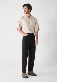 Camisa beige de manga corta con textura vertical, pantalones negros de cintura alta y botines de ante marrón. Silueta simple y relajada.