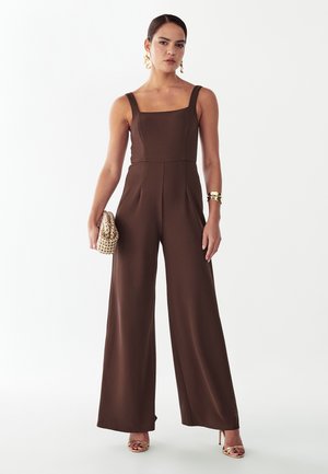 Kobieta stojąca ubrana w brązowy kombinezon bez rękawów z szerokimi nogawkami, trzymająca beżową torebkę z koralikami, z złotymi kolczykami, bransoletką i sandałami na wysokim obcasie.
