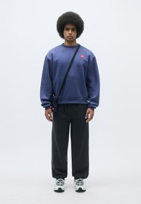 Sudadera morada con un pequeño logo rojo, combinada con pantalones de chándal gris oscuro. El modelo tiene un peinado afrofro y lleva una bolsa cruzada negra.