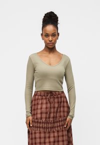 Dugih rukava, maslinasto zeleni crop top rebraste strukture upareni s smeđom kariranom suknjom s elastičnim pojasom i detaljem s vezicama.