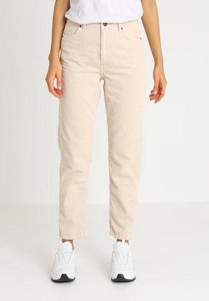 Kvinde iført beige manchesterbukser og hvide sneakers, stående mod en enkel lys baggrund.