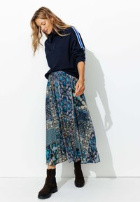 Sweatshirt bleu marin à zip quart avec des rayures blanches, associé à une longue jupe bleue à motifs et des bottines en suède marron.