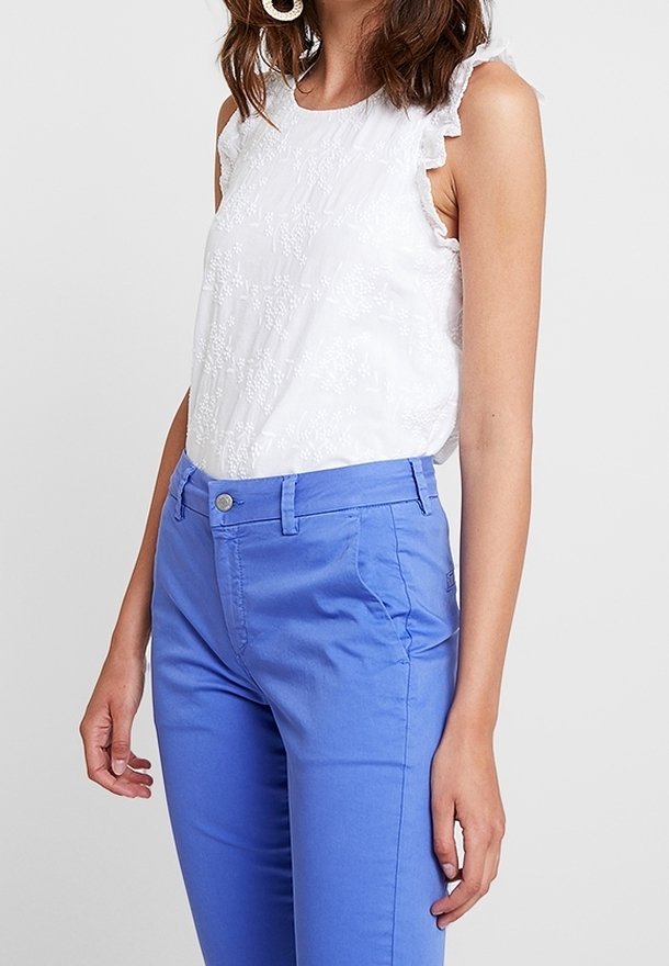 Top bianco senza maniche con dettagli a volant e motivo floreale testurizzato abbinato a pantaloni aderenti blu brillante con tasche frontali e bottone metallico.