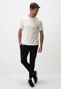 Weißes T-Shirt mit kurzen Ärmeln, kombiniert mit schwarzen, schmal geschnittenen Hosen und schwarzen Sneakers. Das Outfit zeichnet sich durch ein einfaches Design mit klaren Linien aus.