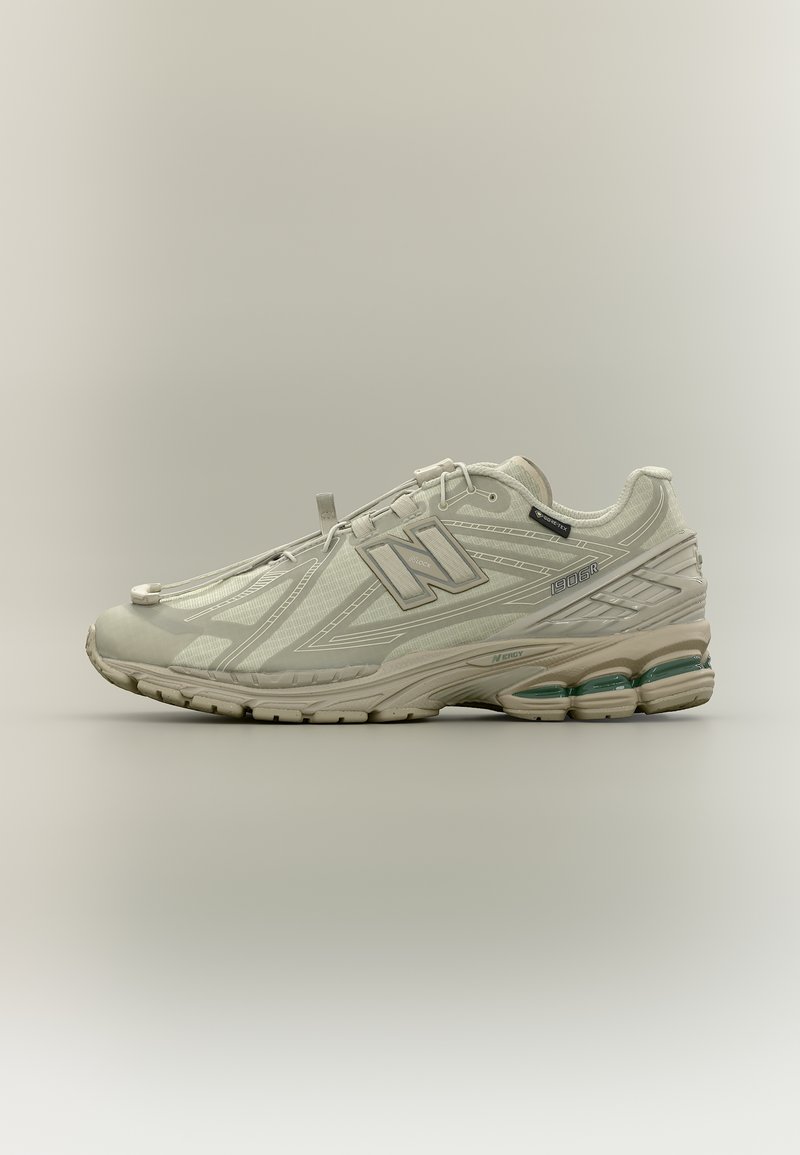 Ανοιχτό γκρι αθλητικό παπούτσι New Balance 1906R με επάνω μέρος από δίχτυ και συνθετικό υλικό, μαλακή σόλα και πράσινες λεπτομέρειες υποστήριξης, εμφανιζόμενο σε πλευρικό προφίλ.