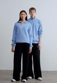 Ανοιχτό μπλε φερμουάρ φούτερ συνδυασμένα με μαύρο παντελόνι με φαρδιά μπατζάκια. Τα φούτερ διαθέτουν μικρό λογότυπο και ριμπ μανσέτες.