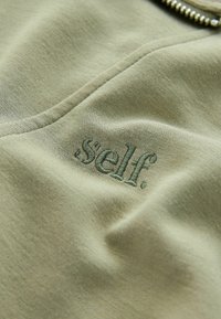 Olivově zelená látka s měkkou texturou, která má výšivku textu ".self." v tmavší zelené barvě, umístěnou ve středu.