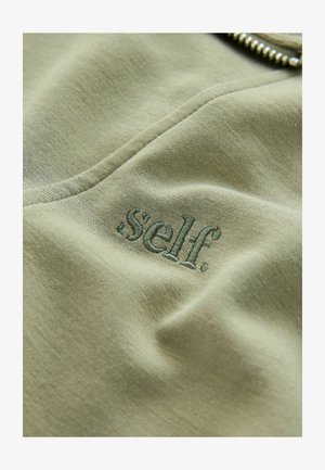 Oliivivihreä kangas, pehmeällä pinnalla, jossa on brodeerattu teksti ".self." tummemmalla vihreällä, sijoitettuna keskelle.