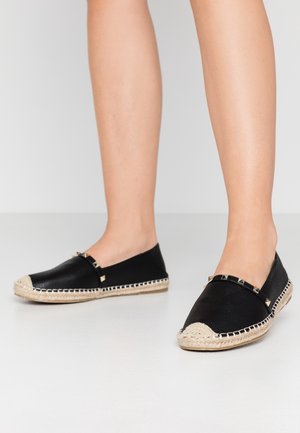 Espadrilles noires avec tige en cuir, semelles plates en jute tissé, bout rond et clous dorés décoratifs sur le devant.