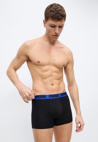 Άνδρας χωρίς πουκάμισο και κοντά μαλλιά ρυθμίζει μαύρα μποξέρ εσώρουχα με μπλε ζωνάκι μέσης με τη μάρκα "PUMA" σε απλό ανοιχτό φόντο.