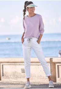 Hellblaues langärmeliges Oberteil, weiße Jogginghosen mit grauem Kordelzug und grauen Sneakers. Visor mit einem schillernden Band. Lässige Strandkulisse.