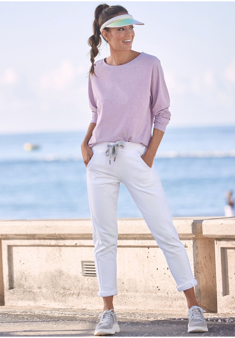 Hellblaues langärmeliges Oberteil, weiße Jogginghosen mit grauem Kordelzug und grauen Sneakers. Visor mit einem schillernden Band. Lässige Strandkulisse.