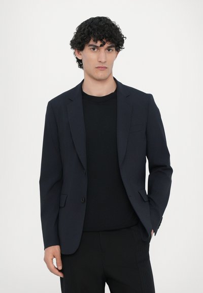 Mørkeblå blazer med slagkraver, enkelt knaplukning og to frontlommer; båret over en sort T-shirt med rund hals. Glat stofstruktur.